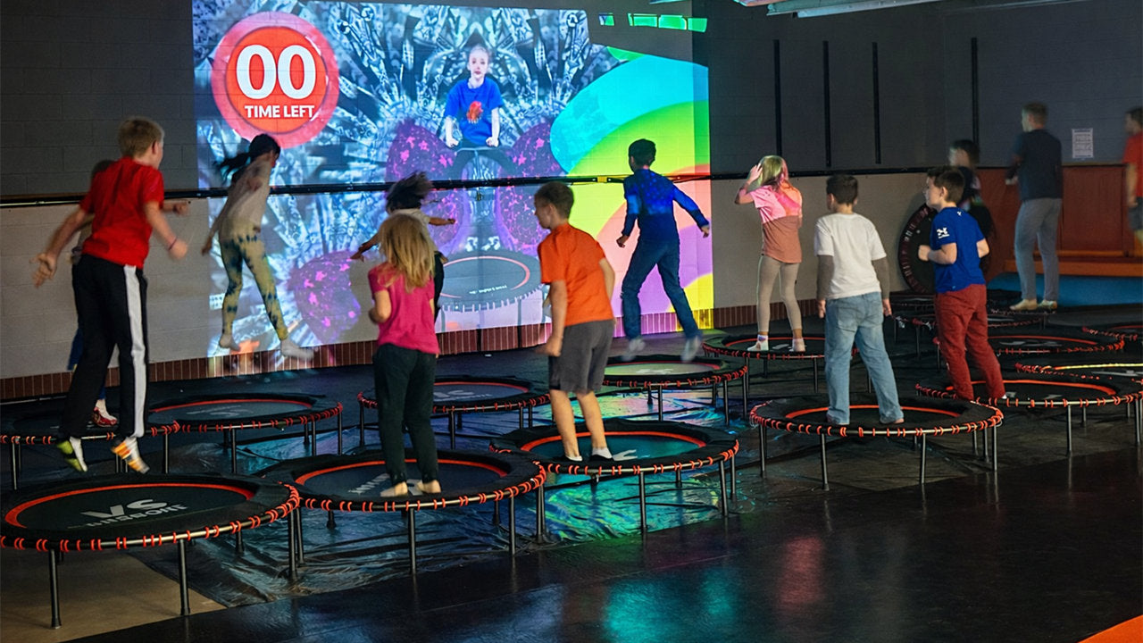 Children playing on trampolines in front of a large screen displaying a game.