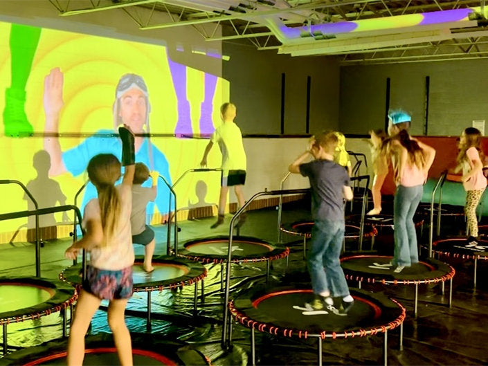Children playing on trampolines in a gymnasium with a large screen displaying colorful graphics.